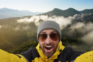 Şapkalı ve güneş gözlüklü gülen adam dağlarda selfie çekiyor.