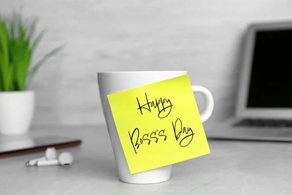 Sticky note with phrase Happy Boss`s Day attached to cup on grey table in office