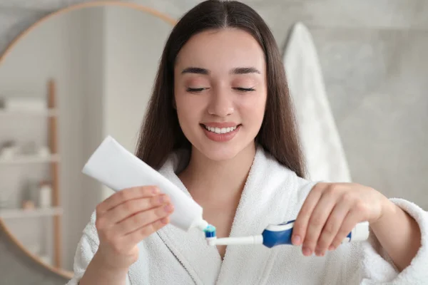 Young woman squeezing toothpaste from tube onto electric toothbrush in bathroom
