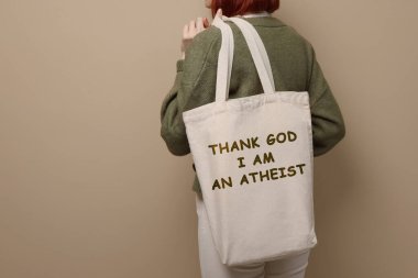 Woman holding bag with phrase Thank God I Am Atheist on beige background, closeup clipart