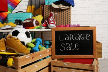 Sign Garage Sale written on blackboard near many different stuff indoors