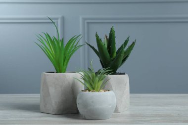 Gri duvarın yanındaki beyaz ahşap masadaki saksılarda birçok farklı yapay bitki var.