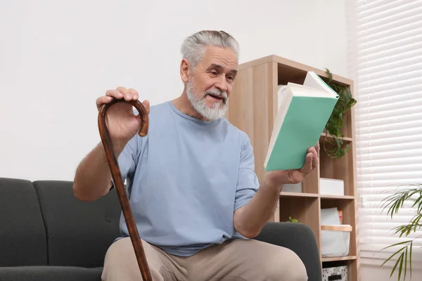 Старший Чоловік Ходячою Тростиною Читає Книгу Дивані Вдома — стокове фото