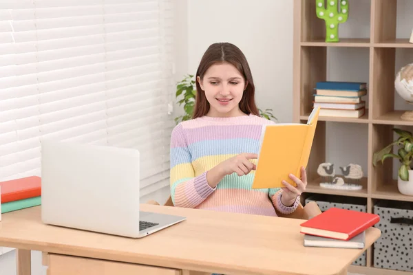 部屋の机でノートパソコンの近くで読書するかわいい女の子 ホームワーク — ストック写真