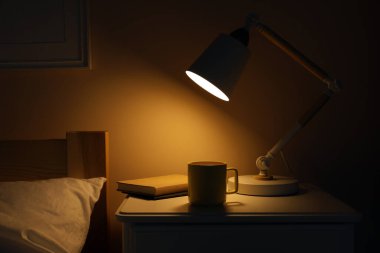 Modern masa lambası, kitaplar ve bir bardak içki karanlık yatak odasındaki beyaz komodinin üzerinde.