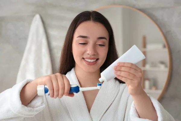 Young woman squeezing toothpaste from tube onto electric toothbrush in bathroom
