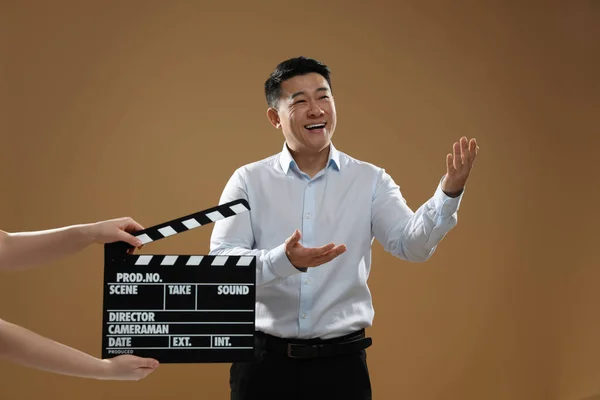 Happy asian actor performing while second assistant camera holding clapperboard on brown background. Film industry