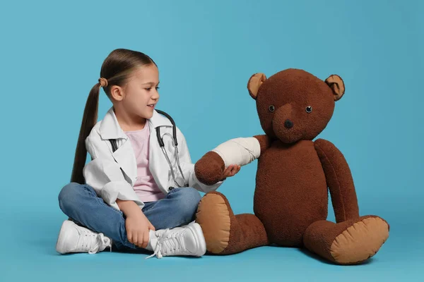 Mała Dziewczynka Mundurze Medycznym Niedźwiedź Zabawka Bandażem Jasnoniebieskim Tle — Zdjęcie stockowe