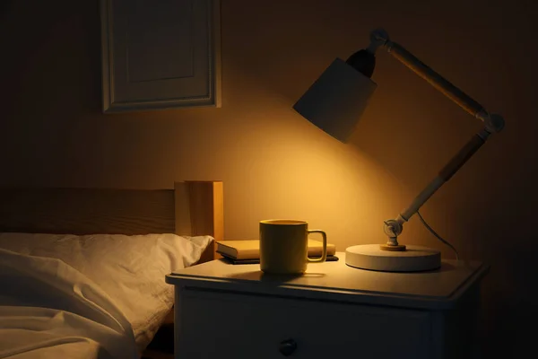 スタイリッシュなモダンなデスクランプ 暗い寝室の白いナイトスタンドでの本と飲み物のカップ — ストック写真