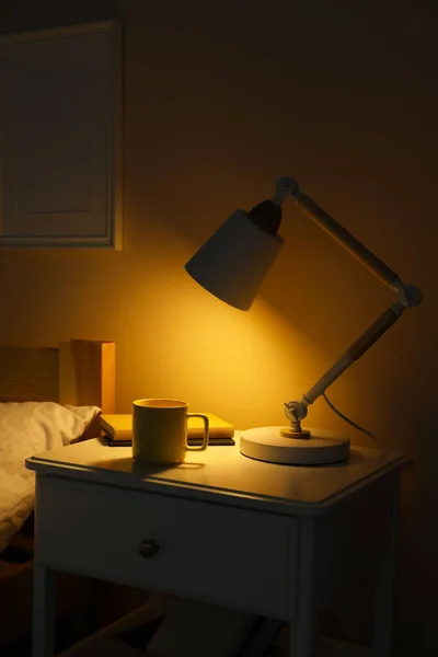 Stylish modern desk lamp, books and cup of drink on white nightstand in dark bedroom