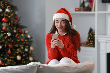 Gülümseyen kadın rahat bir odada kırmızı Noel şapkası ve akıllı telefon takıyor.