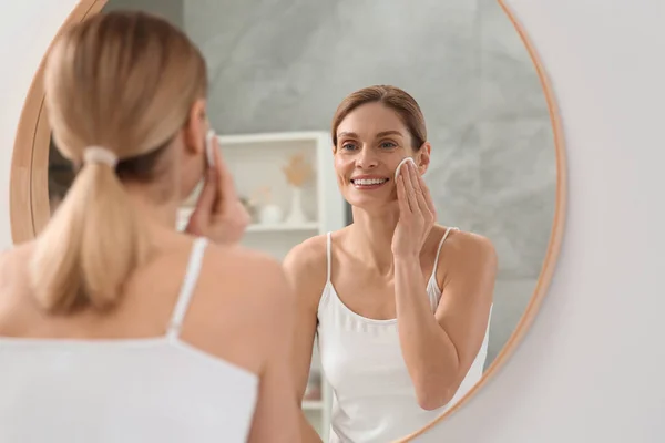 stock image Beautiful woman removing makeup with cotton pad near mirror indoors