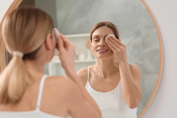 stock image Beautiful woman removing makeup with cotton pad near mirror indoors