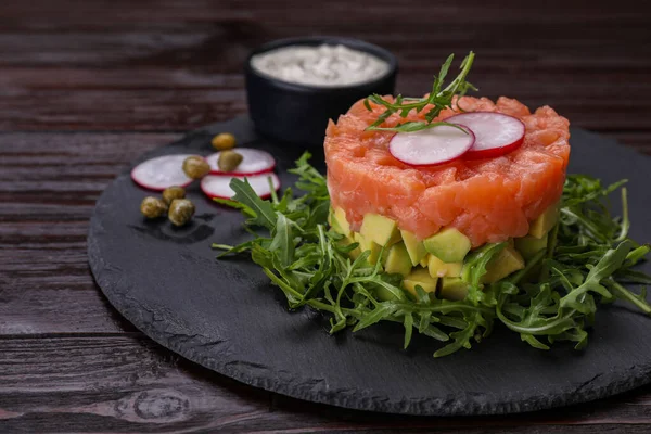 stock image Tasty salmon tartare with radish, avocado and arugula on wooden table, closeup