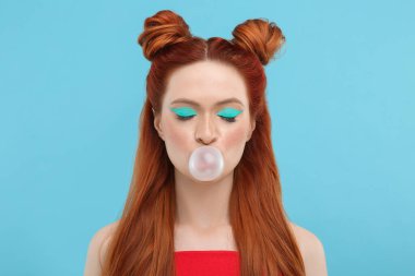 Parlak makyajlı ve kapalı gözlü güzel bir kadın açık mavi arka planda sakız üflüyor.