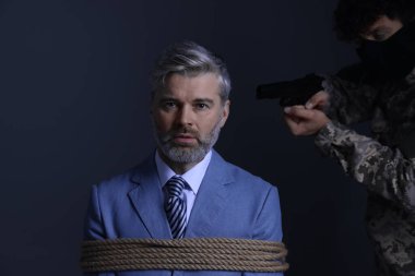 Kidnapper aiming gun at tied victim on dark background. Hostage taking