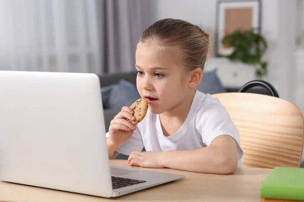 Little girl using laptop and eating biscuit at table indoors. Internet addiction