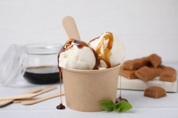 Scoops of ice cream with caramel sauce in paper cup on white tiled table, closeup