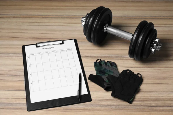 Clipboard with workout plan, dumbbell and cycling gloves on wooden table. Personal training