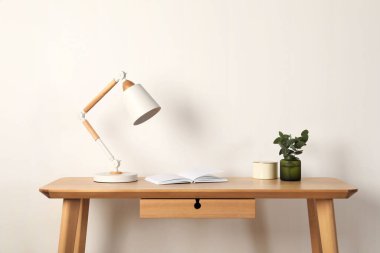 Modern masa lambası, açık kitap ve beyaz duvarın yanındaki ahşap masaya bitki.