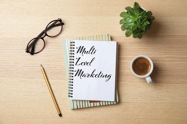 Multi Level Marketing written in notebook, coffee, eyeglasses and houseplant on wooden table, flat lay