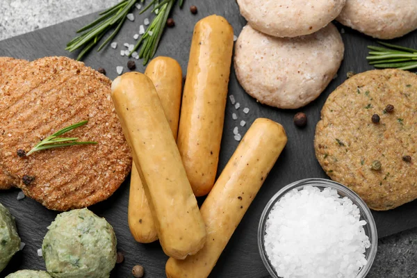 Different raw vegan meat products, salt and rosemary on dark board, flat lay