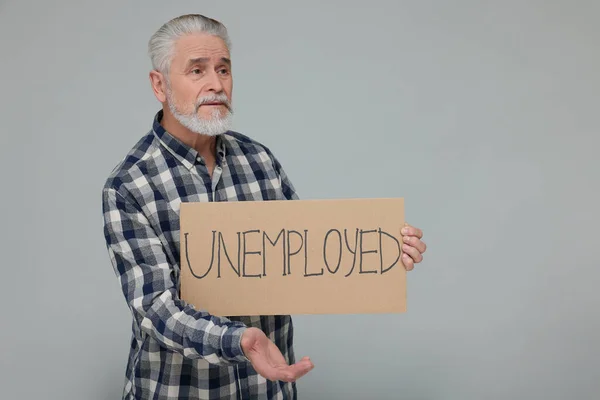 Confused senior man holding cardboard sign with word Unemployed on light grey background. Space for text