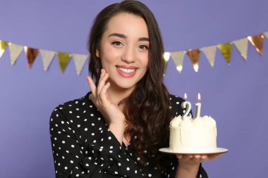 Coming of age party - 21st birthday. Smiling woman holding delicious cake with number shaped candles on violet background