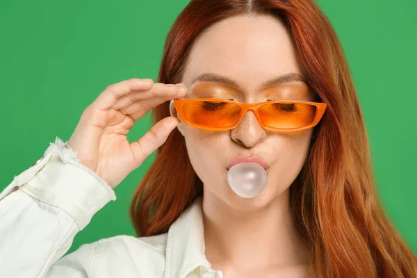 戴着太阳镜 在绿色背景上吹泡泡糖的漂亮女人 — 图库照片