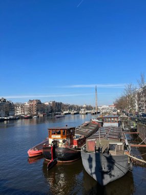 Amsterdam, Hollanda - 01 Mart 2023: Mavi gökyüzü altında demirlemiş teknelerle dolu nehir dolgusu manzarası