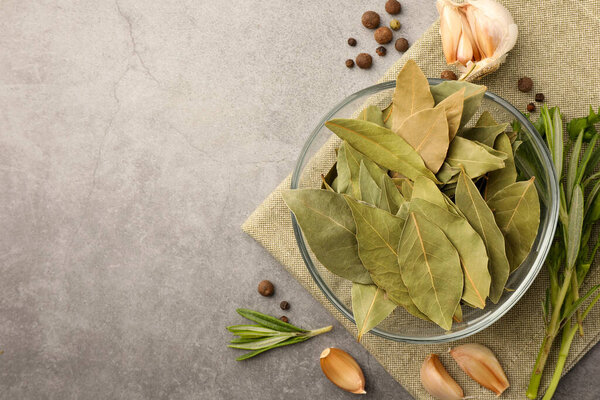 Ароматные лавровые листья в стеклянной миске, различные травы и специи на светлом столе, плоская раскладка. Пространство для текста