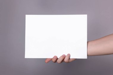 Woman holding sheet of paper on grey background, closeup. Mockup for design