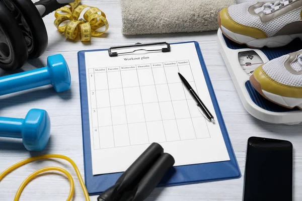 Clipboard with workout plan, smartphone and sports equipment on white wooden table. Personal training