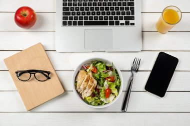Lezzetli yemekler, bir bardak meyve suyu, dizüstü bilgisayar, elma, kitap ve akıllı telefon beyaz ahşap masada, düz yatak. İş yemeği.