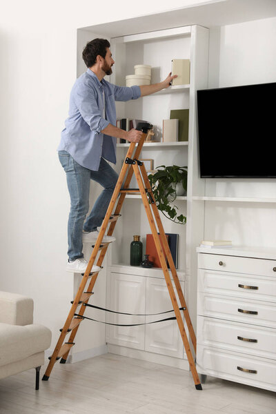Man on wooden folding ladder taking book from shelf at home