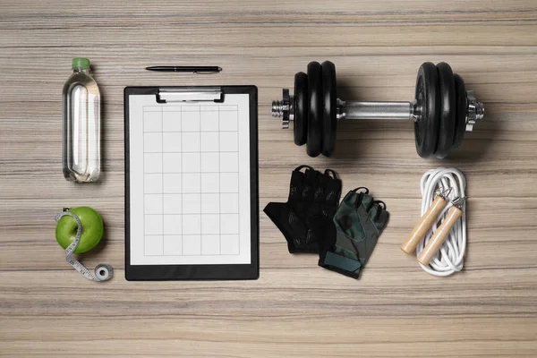 Clipboard with workout plan, apple and sports equipment on wooden table, flat lay. Personal training