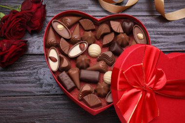 Kalp şeklinde bir kutu, lezzetli çikolata şekerlemeleri, güller ve kurdeleler ahşap masada, düz uzanmış.