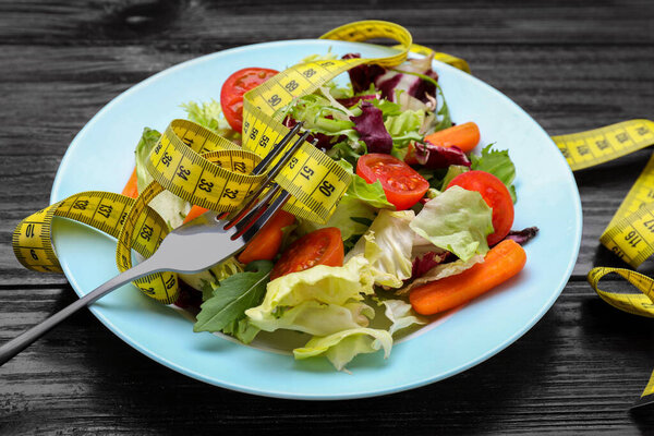 Тарелка со свежим овощным салатом, вилкой и измерительной лентой на деревянном столе, крупным планом. Концепция здорового питания