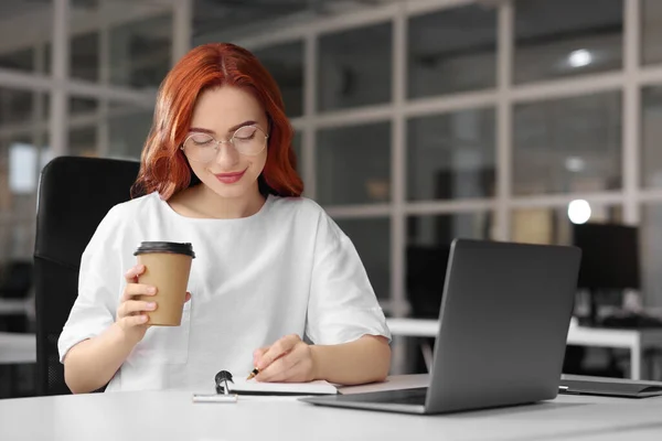 在办公室白桌上的笔记本电脑上工作时 快乐的女人一边喝着纸杯咖啡一边记笔记 — 图库照片