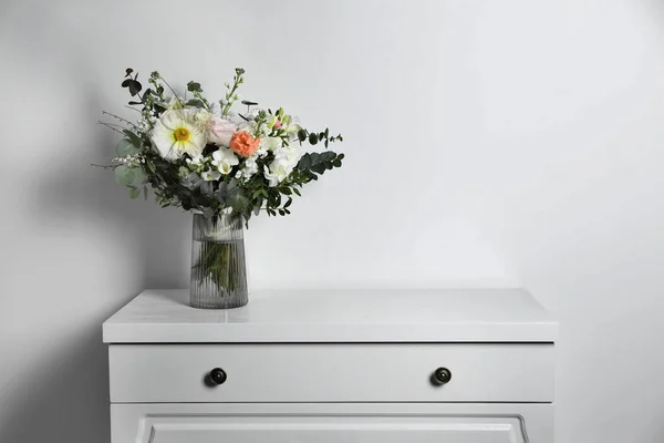 stock image Bouquet with beautiful flowers in vase on chest of drawers near white wall. Space for text