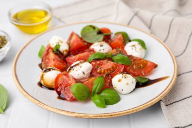 Mozarella topları, domatesler, fesleğen ve sosla beyaz fayanslı masada lezzetli bir Caprese salatası.