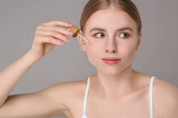 stock image Beautiful young woman applying essential oil onto face on grey background