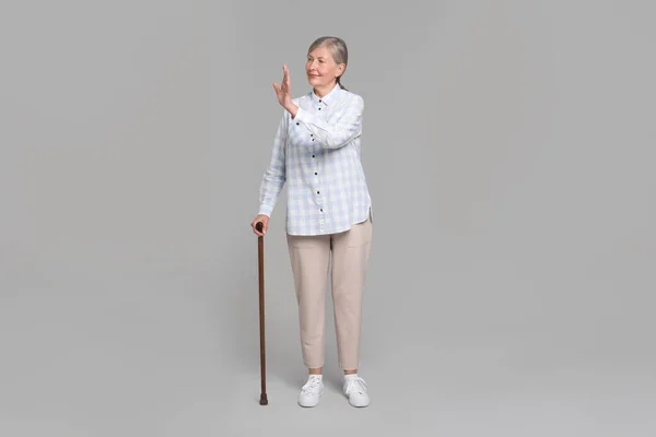 stock image Senior woman with walking cane waving on gray background