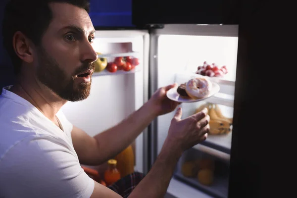 男人在晚上从厨房的冰箱里拿盘子里的甜甜圈 坏习惯 — 图库照片