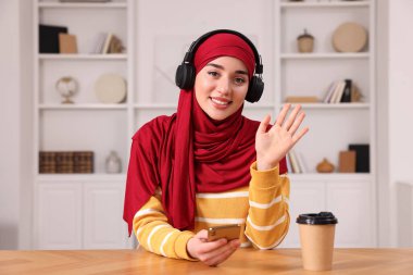 Türbanlı Müslüman kadın odasındaki ahşap masada akıllı telefon kullanıyor.