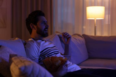 Man eating chips while watching TV on sofa at night. Bad habit