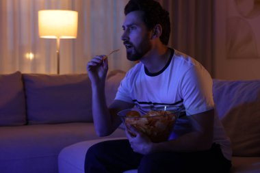 Man eating chips while watching TV on sofa at night. Bad habit