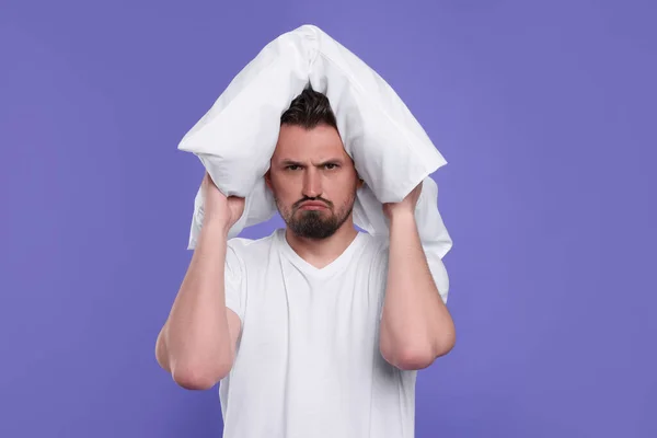 stock image Unhappy man covering ears with pillow on violet background. Insomnia problem