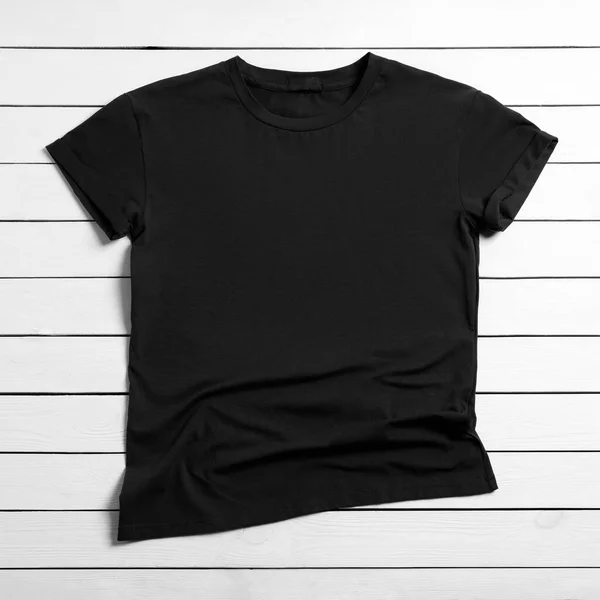 白い木の背景にスタイリッシュな黒のTシャツ トップビュー — ストック写真