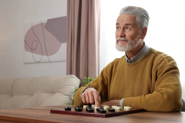 Playing checkers. Senior man thinking about next move at table in room, space for text